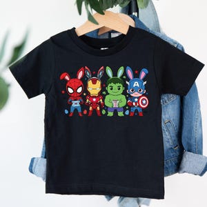 May include: Black t-shirt featuring cartoon versions of Spider-Man, Iron Man, Hulk, and Captain America, each with bunny ears and holding Easter eggs. The shirt is hanging on a wooden hanger against a denim jacket.