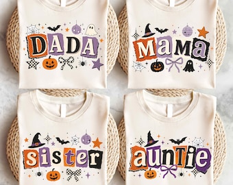 May include: Four cream-colored t-shirts featuring Halloween-themed graphics. Each shirt displays a unique word: DADA, MAMA, sister, or auntie, surrounded by illustrations of pumpkins, ghosts, bats, spiderwebs, disco balls, and bows in orange, purple, and black.