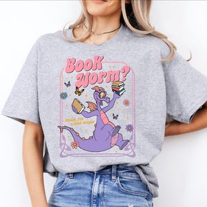 A woman wearing a gray t-shirt with a graphic design featuring a purple dragon holding a book and surrounded by butterflies and flowers.