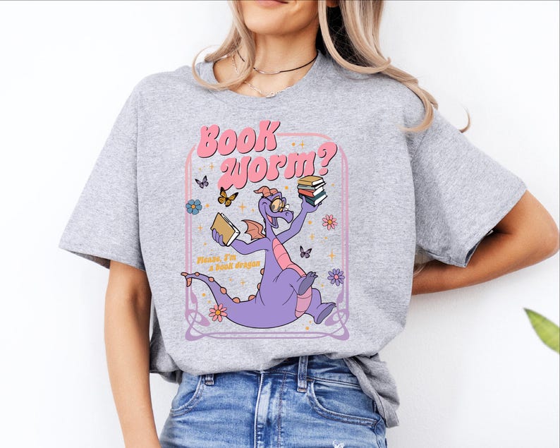 A woman wearing a gray t-shirt with a graphic design featuring a purple dragon holding a book and surrounded by butterflies and flowers.