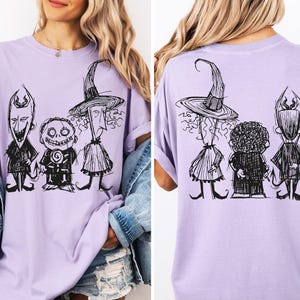May include: A light purple t-shirt featuring a black ink-style sketch graphic of three characters. The front shows the characters facing forward, while the back displays the same three characters from behind, including a tall figure in a pointed hat.