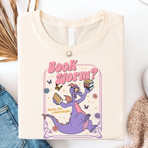 a white t-shirt with a cartoon dragon holding a book, surrounded by butterflies and flowers, along with the text "Book Worm?"