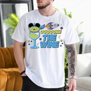 A man wearing a white t-shirt with a graphic design featuring a green alien character and a rocket ship, along with the text "OOOHOH THE WINE".