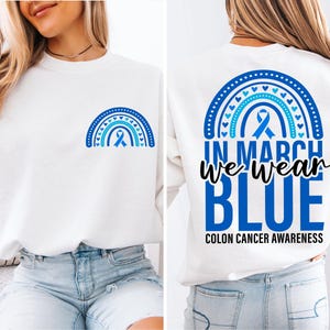 Puede incluir: Sudadera blanca con un diseño de arcoíris de cinta azul en la parte delantera y el texto "In March We Wear Blue Colon Cancer Awareness" en la parte trasera.