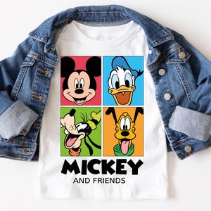 May include: White t-shirt featuring Mickey Mouse, Donald Duck, Goofy, and Pluto in colorful squares. The words "Mickey and Friends" are printed in black below the characters. A denim jacket is draped over the shirt.