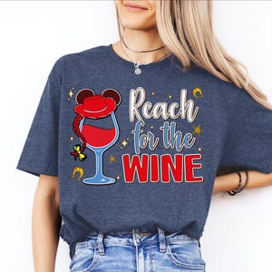 A woman wearing a blue t-shirt with a graphic design that says "Reach for the WINE" and features a wine glass and a character resembling Mickey Mouse.
