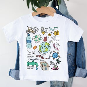 May include: White t-shirt with a colorful graphic promoting environmental awareness. The design includes illustrations of a bicycle, recycling symbols, a globe, and phrases like "Reduce," "Reuse," and "Save the Planet."