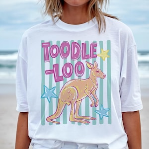 May include: White t-shirt with a kangaroo graphic and the text "TOODLE LOO" in pink. The kangaroo is light orange with yellow accents, set against a background of light green stripes and blue and yellow stars. A casual, fun design.
