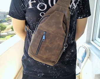 Puede incluir: Un bolso bandolera de cuero marrón con un bolsillo con cremallera y un tirador de cremallera plateado. El bolso se lleva sobre el hombro de una persona que lleva una camisa negra con salpicaduras de pintura blanca.