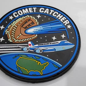 May include: A circular patch featuring a black background with the text COMET CATCHER. It depicts a brown baseball glove catching a blue comet, a light blue and white airplane labeled UNITED STATES OF AMERICA, and a green map of the USA.