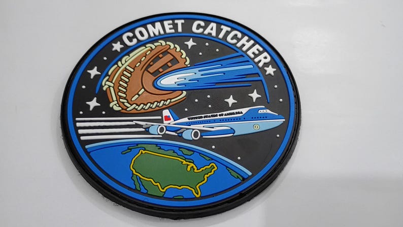 May include: A circular patch featuring a black background with the text COMET CATCHER. It depicts a brown baseball glove catching a blue comet, a light blue and white airplane labeled UNITED STATES OF AMERICA, and a green map of the USA.