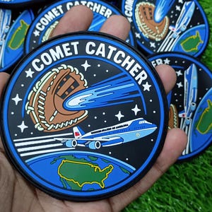 May include: A circular embroidered patch featuring a black background with white stars, a brown baseball glove, a blue comet, and a light blue airplane labeled United States of America flying above a green map of the United States. Text reads COMET CATCHER.