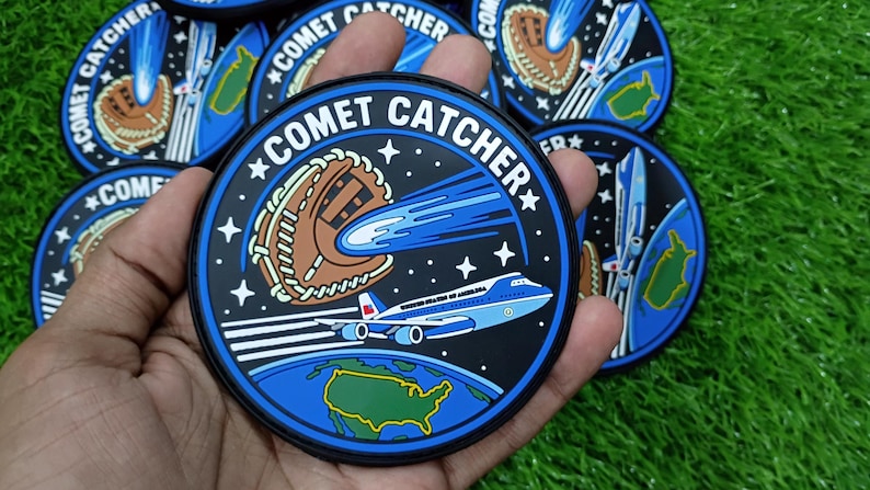 May include: A circular embroidered patch featuring a black background with white stars, a brown baseball glove, a blue comet, and a light blue airplane labeled United States of America flying above a green map of the United States. Text reads COMET CATCHER.