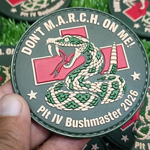 May include: A circular olive green patch featuring a coiled rattlesnake graphic, a red cross symbol, and the text "DON'T M.A.R.C.H. ON ME!" along with "Plt IV Bushmaster 2026" printed in white lettering around the border.