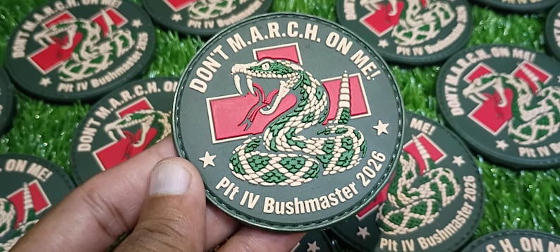 May include: A circular olive green patch featuring a coiled rattlesnake graphic, a red cross symbol, and the text "DON'T M.A.R.C.H. ON ME!" along with "Plt IV Bushmaster 2026" printed in white lettering around the border.
