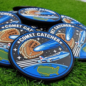 May include: A collection of circular rubber patches featuring the text COMET CATCHER. The design displays a brown baseball glove catching a blue comet above a blue Earth and a white airplane flying over the United States of America.