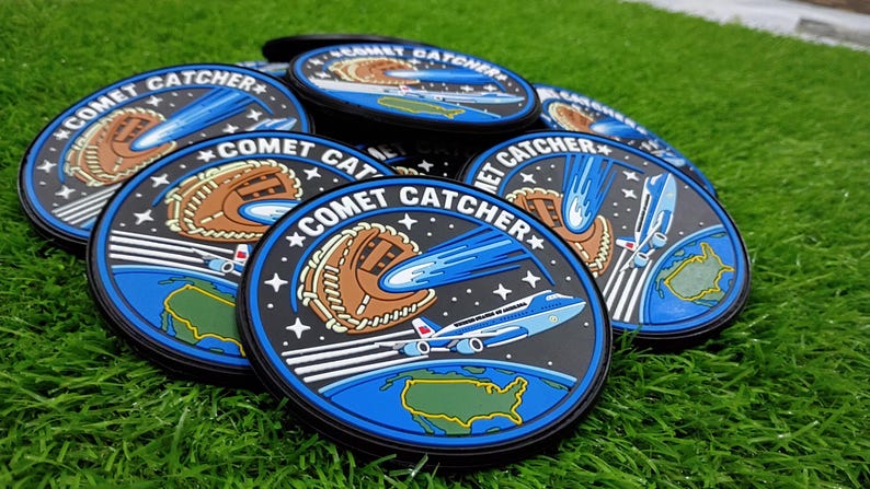 May include: A collection of circular rubber patches featuring the text COMET CATCHER. The design displays a brown baseball glove catching a blue comet above a blue Earth and a white airplane flying over the United States of America.