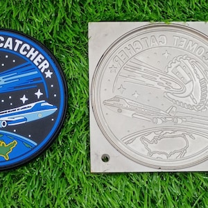 May include: A circular patch and a matching square metal mold featuring the text COMET CATCHER. The design depicts a baseball glove catching a comet, a large airplane, and a map of the United States against a dark blue space background.
