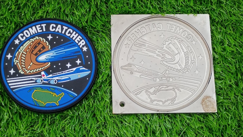 May include: A circular patch and a matching square metal mold featuring the text COMET CATCHER. The design depicts a baseball glove catching a comet, a large airplane, and a map of the United States against a dark blue space background.