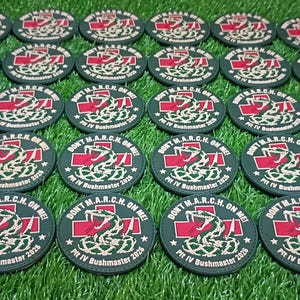May include: A collection of circular black patches featuring a red cross, a white snake graphic, and the text DON'T M.A.R.C.H. ON ME PIT IV BUSHMASTER 2026 arranged in rows on a green surface.