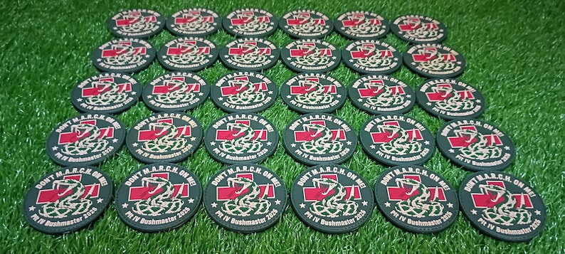 May include: A collection of circular black patches featuring a red cross, a white snake graphic, and the text DON'T M.A.R.C.H. ON ME PIT IV BUSHMASTER 2026 arranged in rows on a green surface.