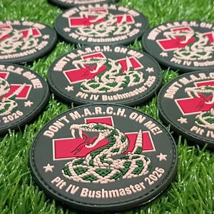 May include: Circular black tactical patches featuring a coiled snake graphic, a red cross, and the text DON'T M.A.R.C.H. ON ME! Plt IV Bushmaster 2026. The design includes white and green details on a dark background with a stitched border.