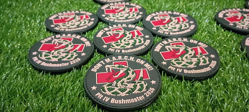 May include: Circular black tactical patches featuring a coiled snake graphic, a red cross, and the text DON'T M.A.R.C.H. ON ME! Plt IV Bushmaster 2026. The design includes white and green details on a dark background with a stitched border.