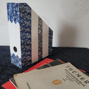 Può includere: Un portariviste verticale decorato con strisce verticali alternate di carta con motivo floreale blu scuro, carta bianca tinta unita e un motivo a stelle geometriche blu e bianco, dotato di un foro circolare per le dita sul pannello frontale.