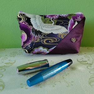 Könnte beinhalten: Eine Kosmetiktasche mit Kranich-Design, mit schwarzem Hintergrund mit goldenen Akzenten und lila Blumenmustern. Die Tasche hat eine tiefviolette Satiseite und einen herzförmigen Silberanhänger. Zwei Kosmetikartikel befinden sich vor der Tasche.