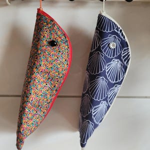 Peut inclure: Deux pochettes en tissu en forme de croissant suspendues à un support. La pochette de gauche présente un motif mosaïque multicolore avec une fermeture éclair rouge et un bouton noir. Celle de droite est bleu marine avec un imprimé coquillages blanc, une fermeture éclair blanche et un bouton transparent.