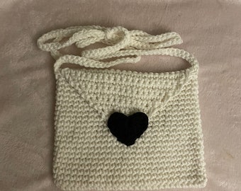 May include: A white crocheted bag with a black heart-shaped embellishment. The bag has a long strap and is perfect for carrying small items.