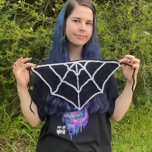 May include: A black and white crocheted spiderweb scarf with a white spiderweb design. The scarf is being held by a person with blue hair and a black t-shirt with a graphic print of a monster.