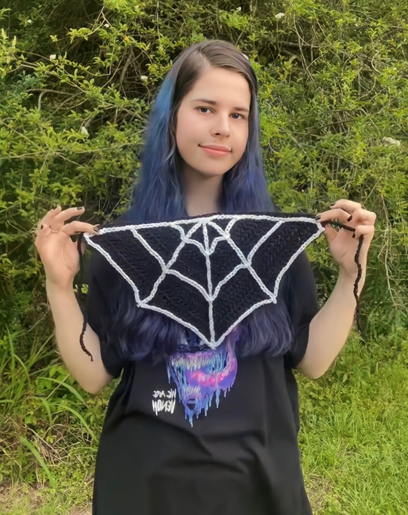 May include: A black and white crocheted spiderweb scarf with a white spiderweb design. The scarf is being held by a person with blue hair and a black t-shirt with a graphic print of a monster.