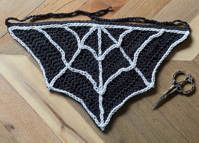 May include: A black and white crocheted spiderweb design with a white border. The web is tied with a black string.