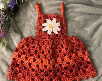 May include: A crocheted dress for a baby or toddler, featuring a white daisy flower on the bodice and a rust-colored ombre design.