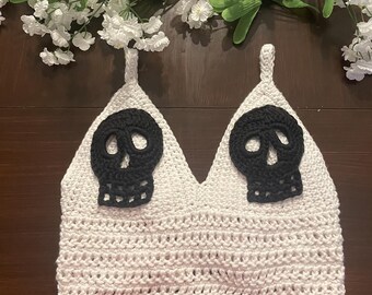 May include: White crochet crop top with black skull appliques. The top has a V-neckline and a scalloped hem.
