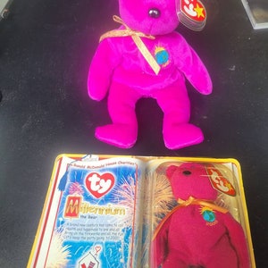 Puede incluir: Un oso de peluche Ty Beanie Baby Millennium de color rosa con una cinta dorada y un globo terráqueo con el año 2000. El oso está sentado sobre un fondo blanco con una etiqueta Ty roja y blanca.