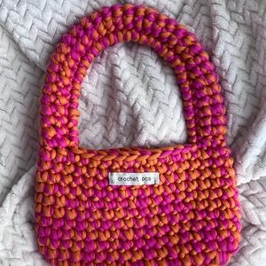 Peut inclure: Un sac crocheté rose et orange avec une petite étiquette indiquant "crochet pcs".