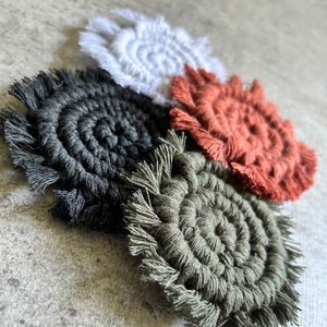 May include: Four handmade macrame coasters in different colors: black, white, green, and orange. Each coaster has a circular shape with a fringe border.