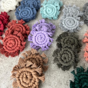 May include: A collection of handmade macrame coasters in various colors, including white, blue, teal, gray, red, orange, purple, brown, olive green, and coral. Each coaster features a circular design with a fringed edge.