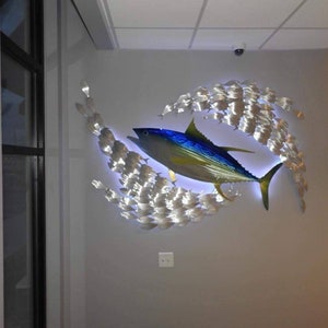 May include: A metal wall sculpture of a blue and yellow fish with a school of smaller silver fish surrounding it. The sculpture is lit from behind, creating a glowing effect.