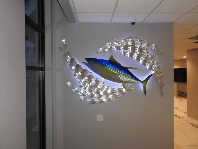 May include: A metal wall sculpture of a blue and yellow fish with a school of smaller silver fish surrounding it. The sculpture is lit from behind, creating a glowing effect.