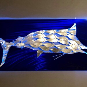 Puede incluir: Una escultura de marlin de metal plateado sobre un fondo azul. El marlin est&aacute; hecho de muchas peque&ntilde;as formas de peces que se superponen. La escultura est&aacute; iluminada con luces LED azules.