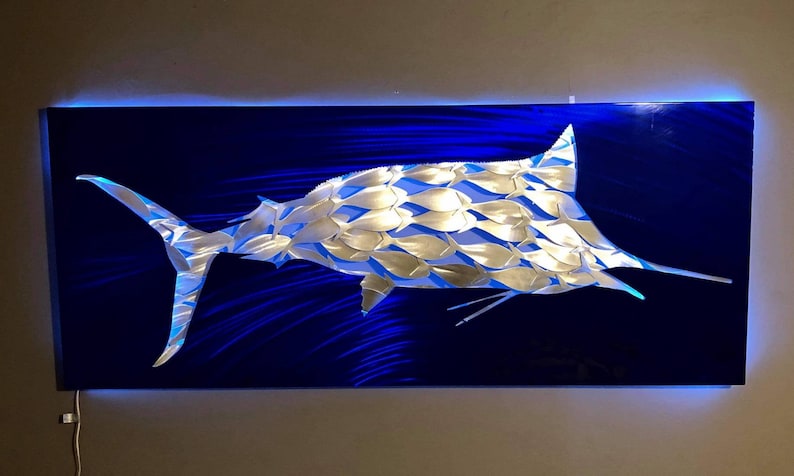 Puede incluir: Una escultura de marlin de metal plateado sobre un fondo azul. El marlin est&aacute; hecho de muchas peque&ntilde;as formas de peces que se superponen. La escultura est&aacute; iluminada con luces LED azules.