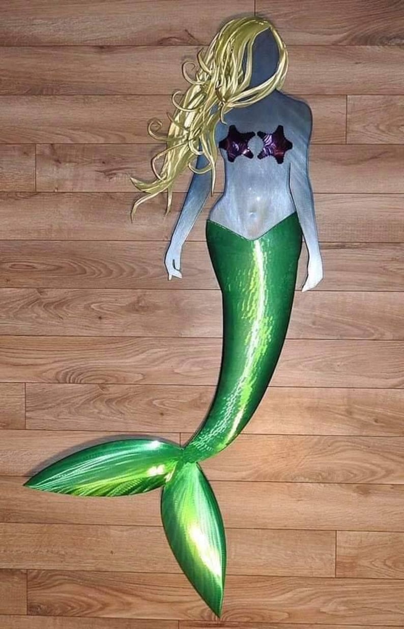 May include: A metal mermaid wall sculpture with a green tail and blonde hair. The mermaid is wearing a seashell bra and has a shiny, metallic finish.