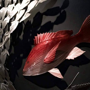 May include: A red fish sculpture with a silver metal seaweed background. The fish is made of metal and has a detailed, realistic design.