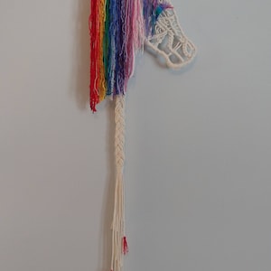 May include: Rainbow yarn macrame wall hanging featuring a unicorn head with a braided tail and colourful tassels. The artwork is signed "MerrittMacrame".