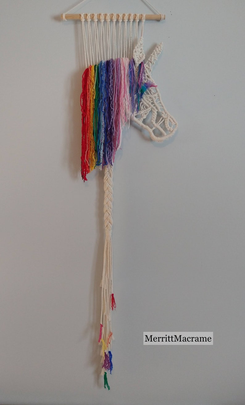 May include: Rainbow yarn macrame wall hanging featuring a unicorn head with a braided tail and colourful tassels. The artwork is signed "MerrittMacrame".