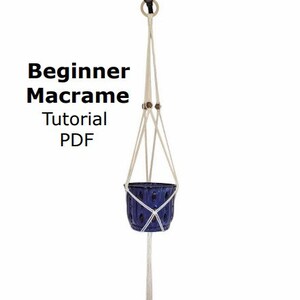 May include: A white macrame plant hanger with a blue ceramic pot. The hanger is made with a simple knot pattern and has a wooden bead at the top. The text "Beginner Macrame Tutorial PDF" is written in black text on a white background.