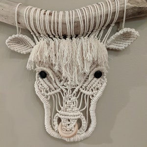May include: A white macrame bull's head wall hanging with black button eyes and a wooden ring nose.